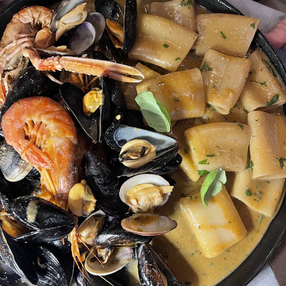 Piatto nero con paccheri in salsa di pesce, cozze, vongole, gamberi e uno scampo, con foglie di basilico.