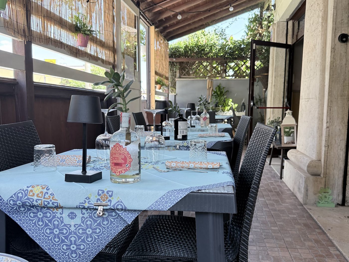 La terrazza coperta del ristorante: tavoli apparecchiati con tovaglie a fantasia maiolica azzurra, sedie in rattan scuro, arelle di canne e piante rampicanti sotto il tetto di travi in legno.