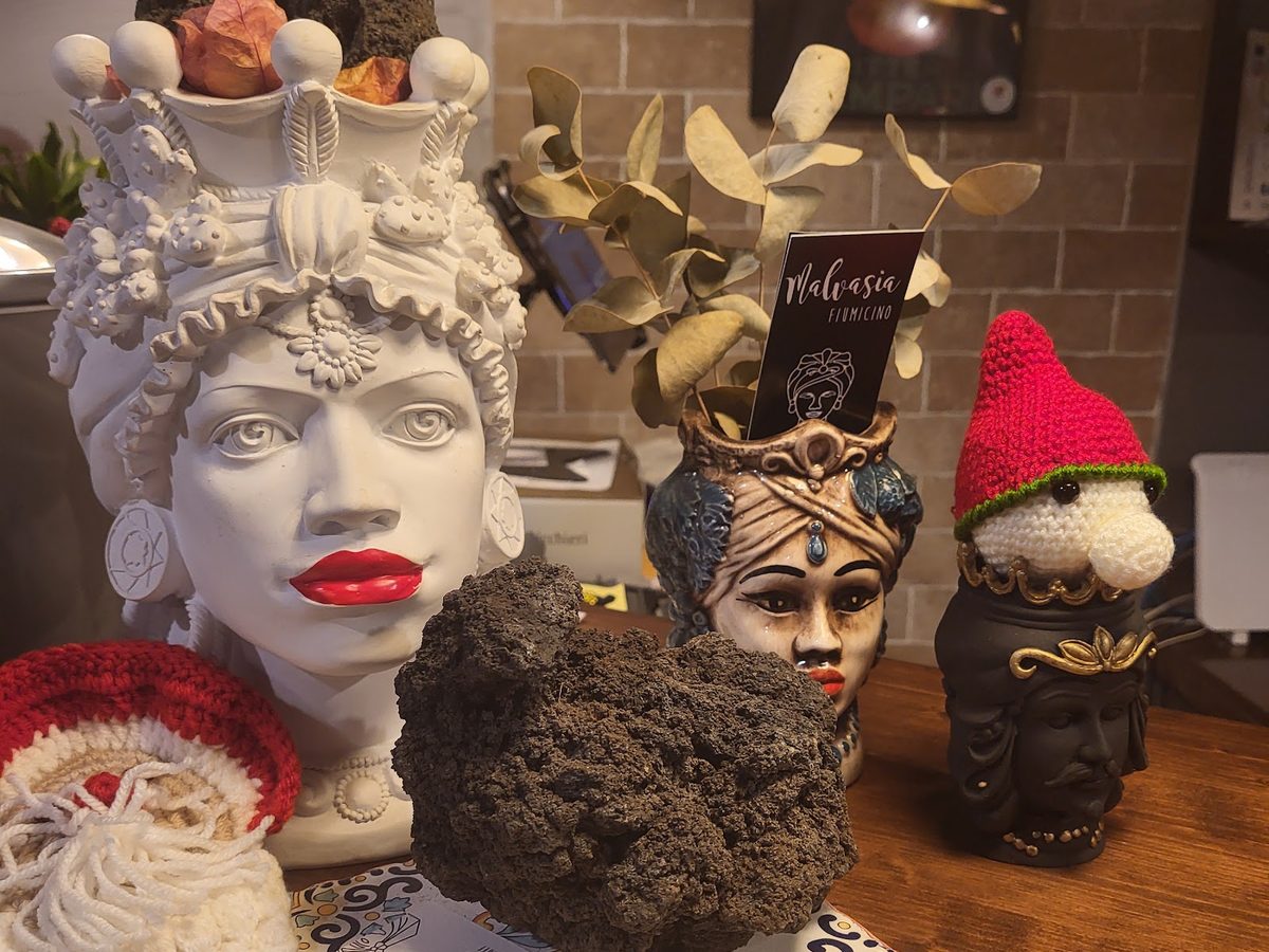 Mensola del locale con teste di moro in ceramica bianca e nera, un blocco di pietra lavica dell&rsquo;Etna, rami di eucalipto e il biglietto da visita del ristorante.